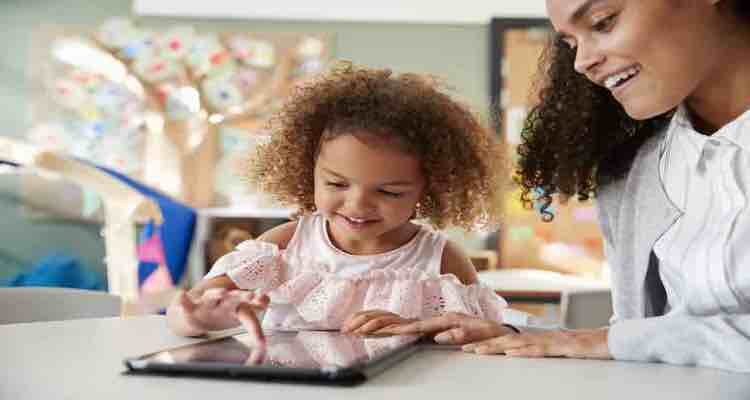 Family Learning Company: Young schoolgirl using a tablet computer with a female infant school teacher, working one on one together in a classroom, close up